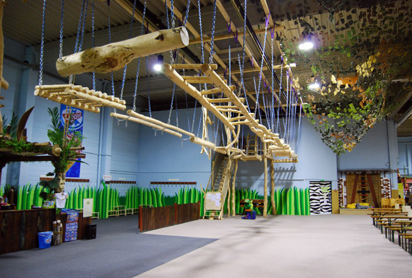 Indoor-Abenteuerpark mit einem hängenden Hindernisparcours aus Holz, grünen Zaundekorationen und verschiedenen Aktivitätsbereichen in einem großen Raum.