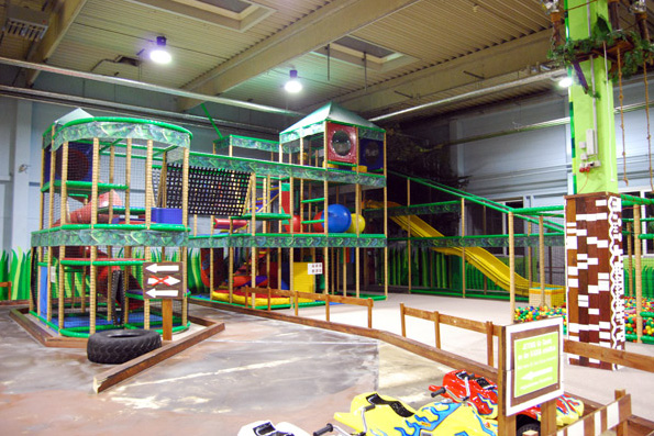 Indoor-Spielplatz mit mehrstufigen Klettergerüsten, Rutschen, Netzen und einem Ballspielplatz, umgeben von Schutzzäunen und heller Deckenbeleuchtung.