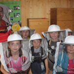 Eine Gruppe von Kindern und ein Erwachsener, die Imkeranzüge und Hüte tragen, stehen in einem Innenraum vor Holzkisten und Lehrplakaten.
