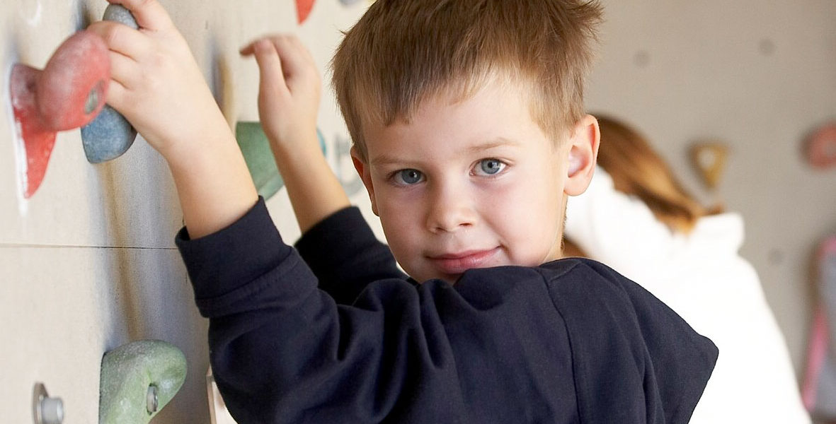 Ein kleiner Junge klettert in einer Halle an einer Felswand und schaut in die Kamera. Deutscher Text wirbt für Kinder- und Jugendferienkletterwochen in einer Salzburger Kletterhalle.