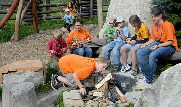 Eine Gruppe von Kindern und ein Erwachsener sitzen um eine Feuerstelle im Freien; ein Kind bläst auf das Feuer, während die anderen Stöcke halten.