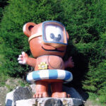 Eine große Cartoon-Bärenstatue mit Schwimmbrille und gestreiftem Schwimmring steht auf Felsen vor dichten grünen Bäumen.