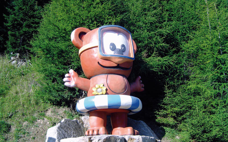 Eine große Cartoon-Bärenstatue mit Schwimmbrille und gestreiftem Schwimmring steht auf Felsen vor dichten grünen Bäumen.