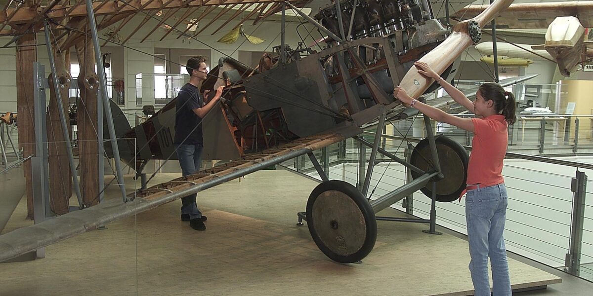 Zwei Personen interagieren mit einem ausgestellten Doppeldecker aus der Zeit des Ersten Weltkriegs; eine Person dreht den Propeller, während die andere das Cockpit untersucht. Der Rahmen und der Motor des Flugzeugs sind ausgestellt.