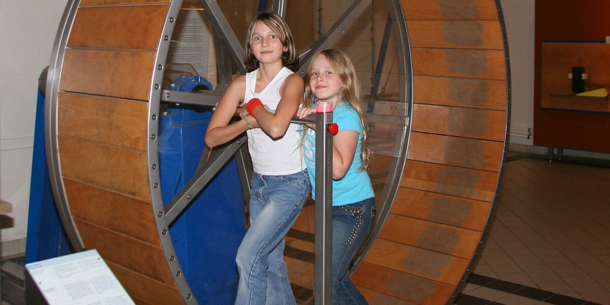 Zwei Mädchen stehen in einem großen hölzernen Hamsterrad in einem Museum, halten sich an Metallstangen fest und schauen in die Kamera.