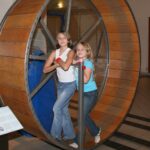 Zwei Mädchen stehen in einem großen hölzernen Hamsterrad in einem Museum, halten sich an Metallstangen fest und schauen in die Kamera.