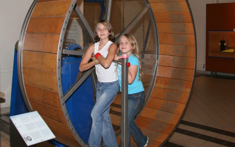 Zwei Mädchen stehen in einem großen hölzernen Hamsterrad in einem Museum, halten sich an Metallstangen fest und schauen in die Kamera.