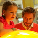 Ein junges Mädchen in einem rosa Kleid und ein Mann in einem roten Pullover beobachten und interagieren mit einem großen leuchtenden gelben Exponat in einem Wissenschaftsmuseum.