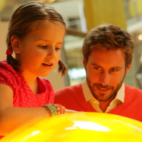 Ein junges Mädchen in einem rosa Kleid und ein Mann in einem roten Pullover beobachten und interagieren mit einem großen leuchtenden gelben Exponat in einem Wissenschaftsmuseum.