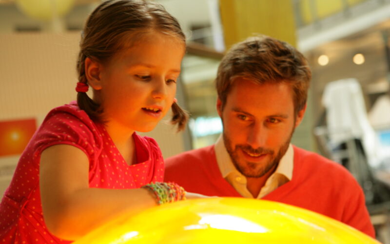 Ein junges Mädchen in einem rosa Kleid und ein Mann in einem roten Pullover beobachten und interagieren mit einem großen leuchtenden gelben Exponat in einem Wissenschaftsmuseum.