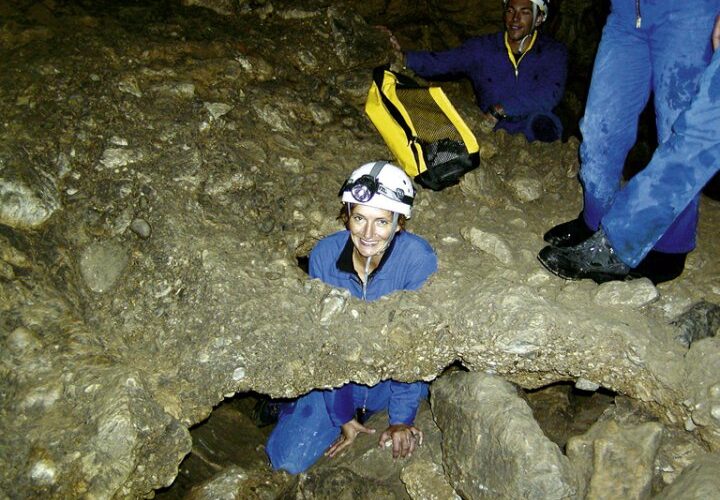 Eine Person in Höhlenausrüstung hockt durch einen engen Felsdurchgang in einer Höhle, während eine andere Person in der Nähe steht, beide tragen Helme mit Stirnlampen.
