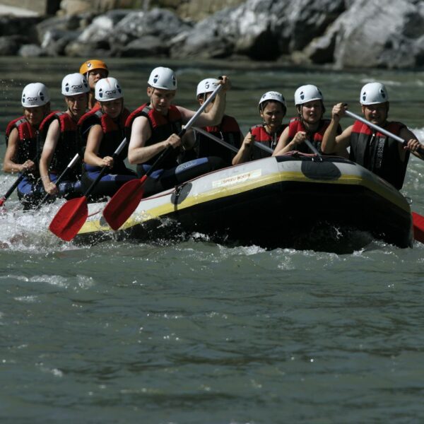 Eine Gruppe von Personen mit Helmen und Schwimmwesten paddelt auf einem Schlauchboot durch einen Fluss mit felsigen Ufern.