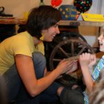Eine Frau und ein kleiner Junge sitzen lächelnd auf dem Boden im Haus. Im Hintergrund sind ein hölzernes Wagenrad und bunte Gegenstände zu sehen.