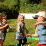 An einem sonnigen Tag spielen mehrere kleine Kinder zusammen auf einer Rasenfläche mit bunten Plastikbällen und im Hintergrund ist ein kleines Zelt zu sehen.