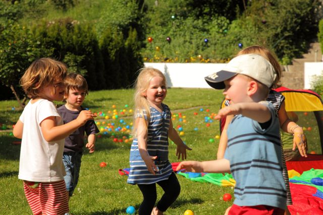 An einem sonnigen Tag spielen mehrere kleine Kinder zusammen auf einer Rasenfläche mit bunten Plastikbällen und im Hintergrund ist ein kleines Zelt zu sehen.
