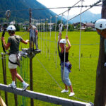 Vier Personen mit Helmen und Klettergurten navigieren in einem Seilgarten im Freien, während im Hintergrund Berge und Gebäude zu sehen sind.