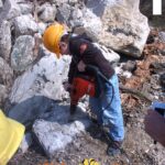 Ein Kind mit einem gelben Schutzhelm arbeitet mit einer Bohrmaschine an einem großen Felsen im Freien, mit Schutt im Hintergrund. Der Text "Kids on Stage" ist am unteren Rand eingeblendet.