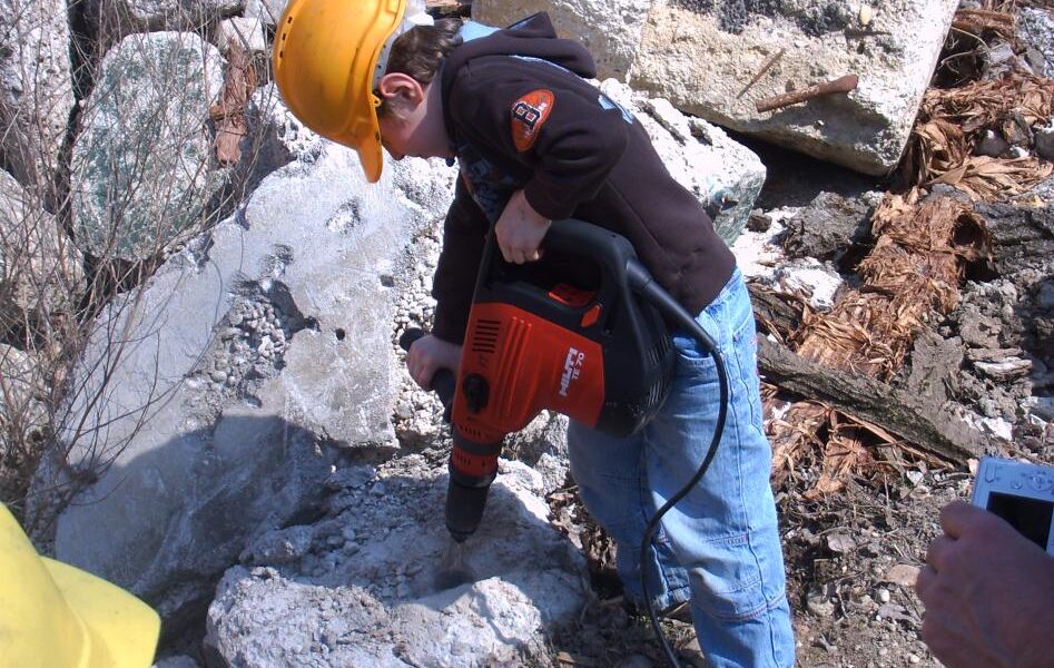 Ein Kind mit einem gelben Schutzhelm arbeitet mit einer Bohrmaschine an einem großen Felsen im Freien, mit Schutt im Hintergrund. Der Text "Kids on Stage" ist am unteren Rand eingeblendet.
