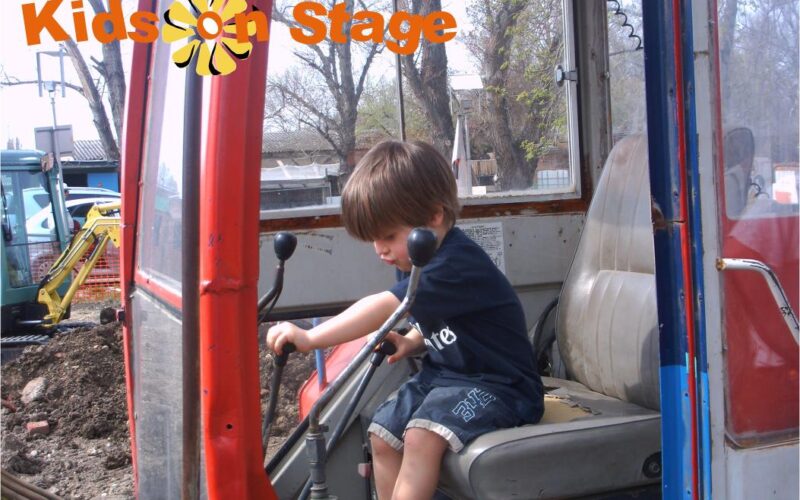 Ein kleines Kind sitzt auf dem Fahrersitz einer Baumaschine und hält die Steuerung in der Hand. Oben ist der Text "Kids on Stage" eingeblendet. In der Ecke erscheint das Datum "2009-04-05".