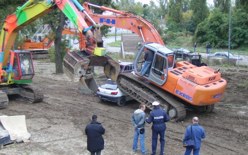 Zwei Bagger zertrümmern ein Auto, während mehrere Personen, darunter zwei Polizisten, die Szene auf einem von Bäumen umgebenen Grundstück beobachten.