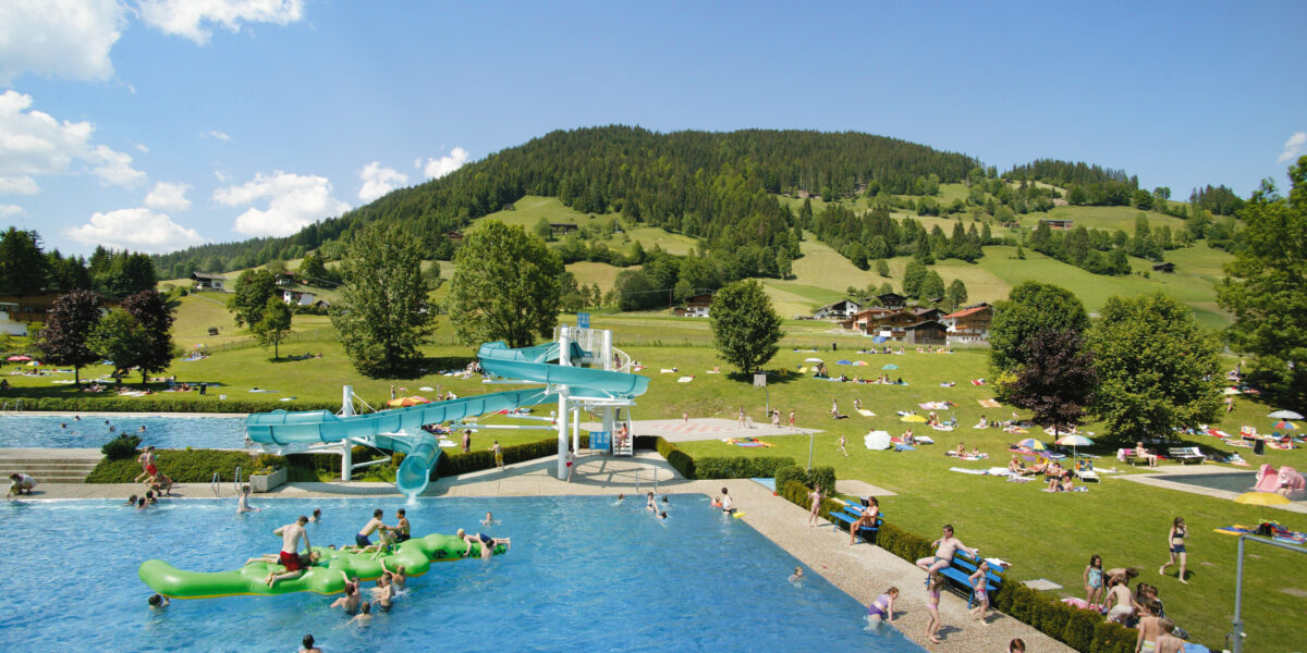 Freibad mit schwimmenden und spielenden Menschen, einer Wasserrutsche und grasbewachsenen Hügeln mit Bergen und verstreuten Sonnenanbetern im Hintergrund an einem sonnigen Tag.