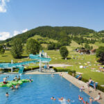 Freibad mit schwimmenden und spielenden Menschen, einer Wasserrutsche und grasbewachsenen Hügeln mit Bergen und verstreuten Sonnenanbetern im Hintergrund an einem sonnigen Tag.