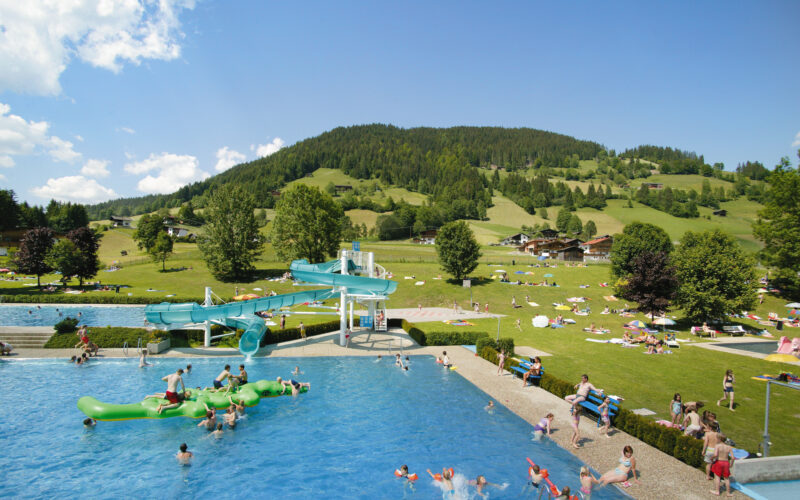 Freibad mit schwimmenden und spielenden Menschen, einer Wasserrutsche und grasbewachsenen Hügeln mit Bergen und verstreuten Sonnenanbetern im Hintergrund an einem sonnigen Tag.