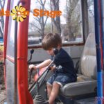Ein kleines Kind bedient die Steuerelemente in der Kabine eines Baufahrzeugs. Oben ist der Text "Kids on Stage" eingeblendet, unten rechts steht das Datum "2009-04-05".