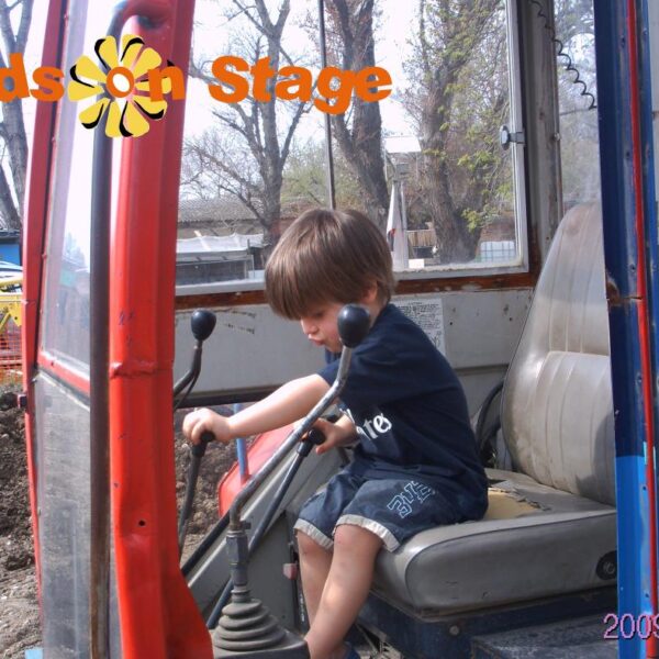 Ein kleines Kind bedient die Steuerelemente in der Kabine eines Baufahrzeugs. Oben ist der Text "Kids on Stage" eingeblendet, unten rechts steht das Datum "2009-04-05".