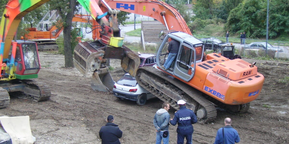 Zwei Bagger, ein bunt lackierter und ein orangefarbener, sind auf dem Boden geparkt; der orangefarbene Bagger zertrümmert gerade ein Auto. Vier Personen, darunter zwei Polizisten, stehen in der Nähe und beobachten.