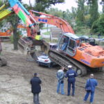 Zwei Bagger, ein bunt lackierter und ein orangefarbener, sind auf dem Boden geparkt; der orangefarbene Bagger zertrümmert gerade ein Auto. Vier Personen, darunter zwei Polizisten, stehen in der Nähe und beobachten.