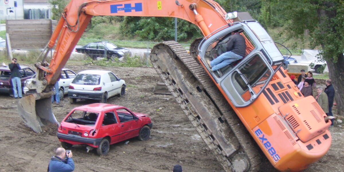 Ein Bagger hebt sein vorderes Ende an, indem er seine Schaufel auf ein rotes Auto stellt, während mehrere Personen in der Nähe stehen, beobachten und Fotos machen.