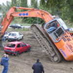 Ein Bagger hebt sein vorderes Ende an, indem er seine Schaufel auf ein rotes Auto stellt, während mehrere Personen in der Nähe stehen, beobachten und Fotos machen.