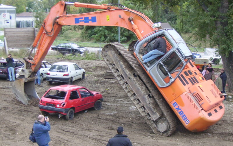Ein Bagger hebt sein vorderes Ende an, indem er seine Schaufel auf ein rotes Auto stellt, während mehrere Personen in der Nähe stehen, beobachten und Fotos machen.