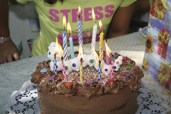 Ein Schokoladengeburtstagskuchen mit brennenden Kerzen und Süßigkeiten steht auf einem Tisch; im Hintergrund ist eine Person in einem gelben Hemd zu sehen.