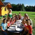 Eine Gruppe von Kindern und zwei Erwachsene sitzen an einem langen Tisch im Freien, der für eine Mahlzeit in der Nähe eines Holzgebäudes an einem sonnigen Tag gedeckt ist.