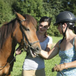 Zwei Frauen stehen neben einem braunen Pferd auf einer grasbewachsenen Wiese; eine Frau streichelt das Pferd, während die andere lächelnd zusieht. Ein anderes Pferd ist teilweise rechts zu sehen.