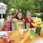 Drei Mädchen sitzen an einem mit gelben und orangefarbenen Gegenständen dekorierten Tisch im Freien, lächeln und machen sich gegenseitig Hasenohren hinter dem Kopf. Ein gelbes Blumenplüschtier liegt auf dem Tisch.