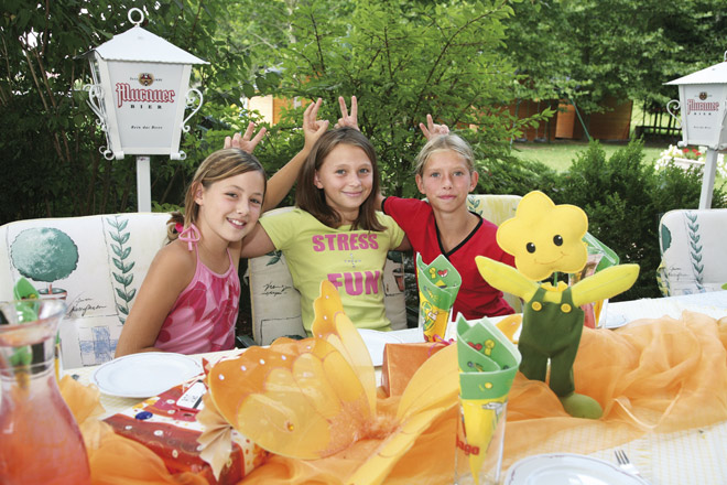Drei Mädchen sitzen an einem mit gelben und orangefarbenen Gegenständen dekorierten Tisch im Freien, lächeln und machen sich gegenseitig Hasenohren hinter dem Kopf. Ein gelbes Blumenplüschtier liegt auf dem Tisch.