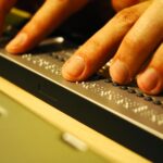 Nahaufnahme von Händen, die Blindenschrift auf einer aktualisierbaren Braillezeile neben einer Laptop-Tastatur lesen.