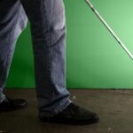 Person in Jeans und schwarzen Schuhen, die mit einem weißen Stock auf einem Betonboden geht, vor einem grünen Hintergrund stehend.