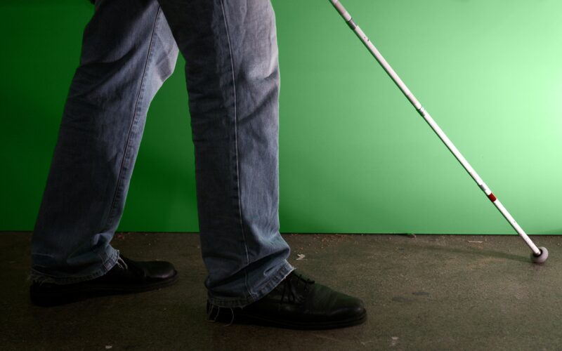 Person in Jeans und schwarzen Schuhen, die mit einem weißen Stock auf einem Betonboden geht, vor einem grünen Hintergrund stehend.