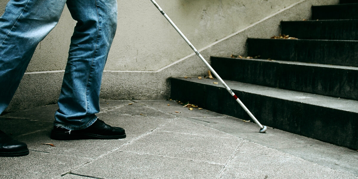 Eine Person in Jeans und schwarzen Schuhen benutzt einen weißen Stock, um sich in der Nähe einer Treppe auf einem Betonweg zurechtzufinden.