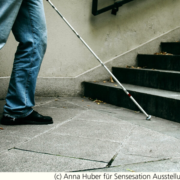 Eine Person in Jeans und schwarzen Schuhen benutzt einen weißen Stock, um sich in der Nähe einer Treppe auf einem Betonweg zurechtzufinden.
