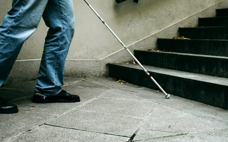 Eine Person in Jeans und schwarzen Schuhen benutzt einen weißen Stock, um sich in der Nähe einer Treppe auf einem Betonweg zurechtzufinden.