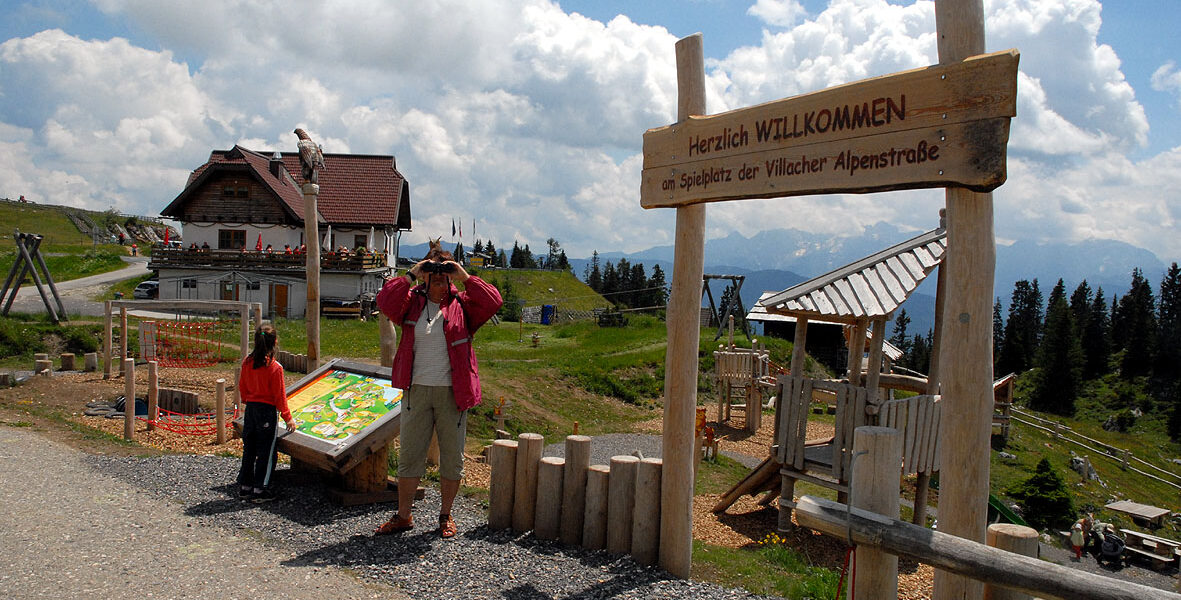 Ein Mann macht ein Foto in der Nähe eines hölzernen Willkommensschildes an einem Aussichtspunkt in den Bergen, mit einem Kind und einem Gebäude im Chalet-Stil im Hintergrund unter einem teilweise bewölkten Himmel.