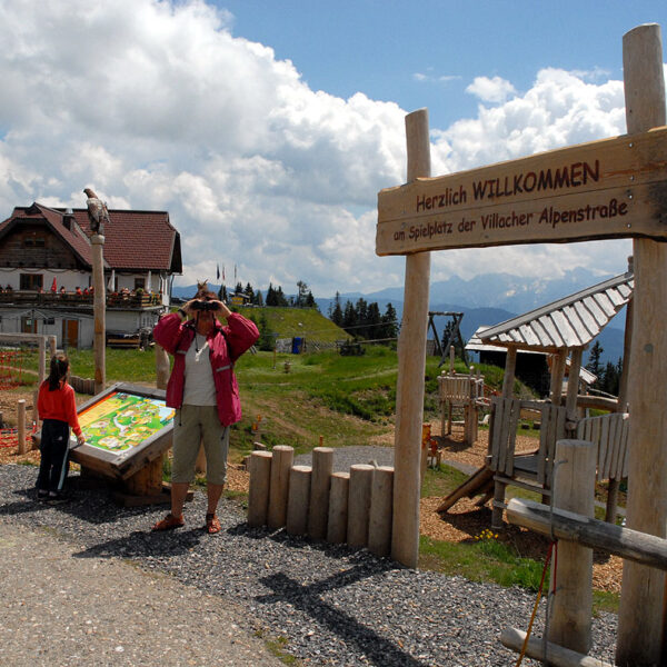 Ein Mann macht ein Foto in der Nähe eines hölzernen Willkommensschildes an einem Aussichtspunkt in den Bergen, mit einem Kind und einem Gebäude im Chalet-Stil im Hintergrund unter einem teilweise bewölkten Himmel.