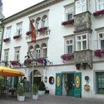 Ein weißes Gebäude mit Blumenkästen und Bogenfenstern, das als "Hotel Post" bezeichnet wird, mit Sitzgelegenheiten im Freien und Menschen in der Nähe.