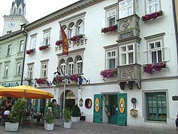 Ein weißes Gebäude mit Blumenkästen und Bogenfenstern, das als "Hotel Post" bezeichnet wird, mit Sitzgelegenheiten im Freien und Menschen in der Nähe.