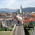 Eine Brücke führt über einen Fluss zu einer Stadt mit bunten Gebäuden und einem hohen Kirchturm vor der Kulisse der Berge.